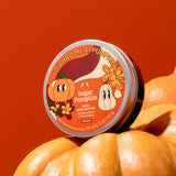 Container of The Body Shop's Sugar Pumpkin body butter sitting on large pumpkins with a red background